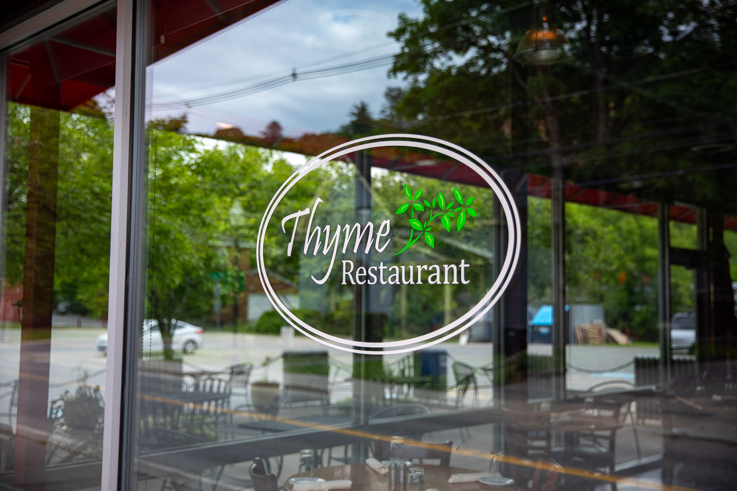 005A7505 A glass window with the logo for Thyme Restaurant, featuring elegant white text and a green herb graphic, with reflections of outdoor seating and trees in the background.