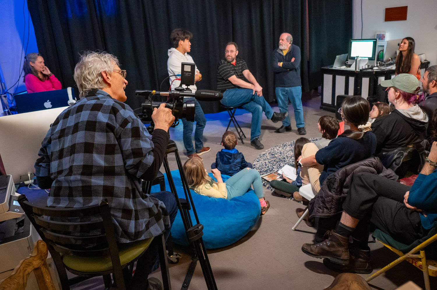 A group of three men stand at the front of a room giving a presentation to a small group of spectators. To the left a camera man sits, recording the event. 