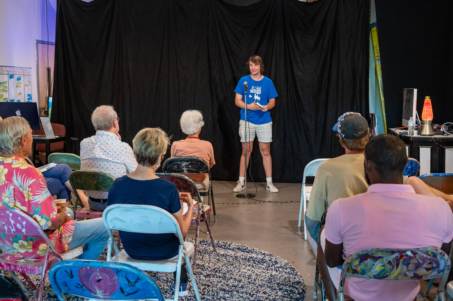 A young man stands on a small stage in front of a black curtain, speaking into a microphone while holding a piece of paper. He wears a blue t-shirt with a media logo, khaki shorts, and sneakers. An audience of older adults sits in colorful mismatched chairs, listening attentively. The setting is casual and creative, with a lava lamp and electronics visible on a side table, giving the space a community arts vibe.
